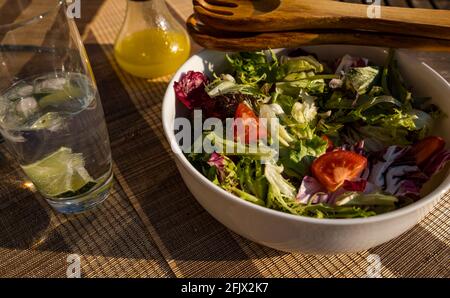 Bol à salade avec légumes verts, laitue et tomates, salade en bois, cuillères à servir sur une table extérieure au soleil, avec une bouteille de vinaigrette Banque D'Images