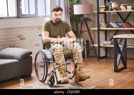 Jeune vétéran handicapé barbu assis dans un fauteuil roulant et regardant à l'appareil photo Banque D'Images