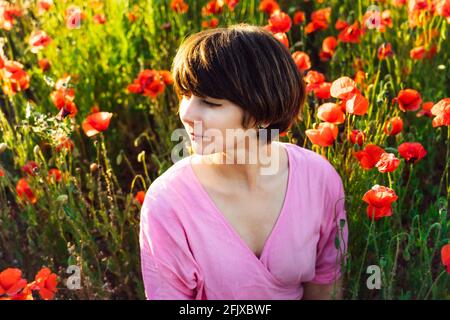 Gros plan mixte race femme en robe rose se détendre dans rouge coquelicots fleurs prairie dans la lumière du coucher du soleil. Simple plaisir pour la santé mentale et le bien-être Banque D'Images