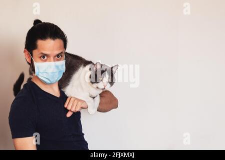 Jeune latino avec un chat sur ses épaules portant Un masque de sécurité de COVID-19 sur fond clair avec espace de copie Banque D'Images