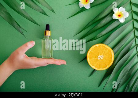 Sérum cosmétique vitamine C en flacon de verre avec pipette compte-gouttes dans la main femelle. Huile essentielle d'orange avec de la vitamine C d'agrumes et des feuilles de palmier sur la couleur Banque D'Images