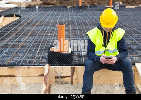 Un ouvrier de la construction parle sur un smartphone dans un casque jaune et gilet de protection sur le fond de la construction de renforcement de la maison pour Banque D'Images