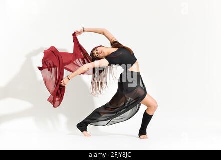 Danse gracieuse effectuant une pose de patinage artistique Ina Bauer un costume noir filmy flottant un rectangle de tissu rouge derrière elle sur blanc avec ombre Banque D'Images