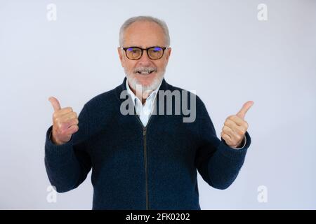 Homme senior aux cheveux gris debout sur un fond bleu isolé faisant un geste de pouce heureux vers le haut avec la main. Approbation de l'expression en regardant la caméra showi Banque D'Images