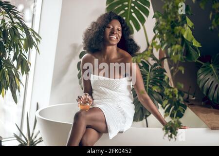 Femme souriante à la peau sombre assise dans la salle de bains et se sentant détendue Banque D'Images