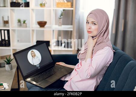 Fatiguée jeune femme musulmane fatiguée atteinte d'un mal de gorge, appelant à une conversation vidéo avec une femme médecin via l'application de soins de santé, assise sur un canapé à la maison avec un ordinateur portable. Télémédecine, concept de technologie Banque D'Images