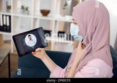 Pandémie de coronavirus. Femme musulmane malade dans le hijab et le masque, souffrant de mal de gorge, utilisant un Tablet pc et de passer un appel vidéo avec un médecin indien mâle tout en restant à la maison Banque D'Images