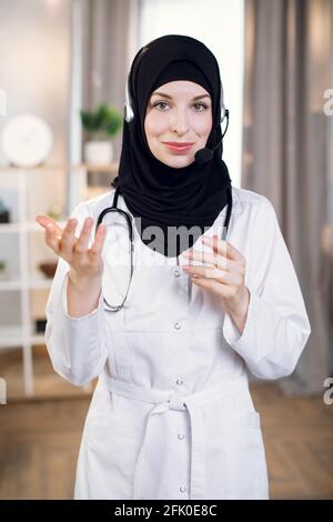 Agréable femme musulmane médecin portant un foulard noir, utilisant un casque, gestante et parlant à la caméra, tout en travaillant à la clinique. Femme médecin généraliste arabe ayant un rendez-vous en ligne. Banque D'Images