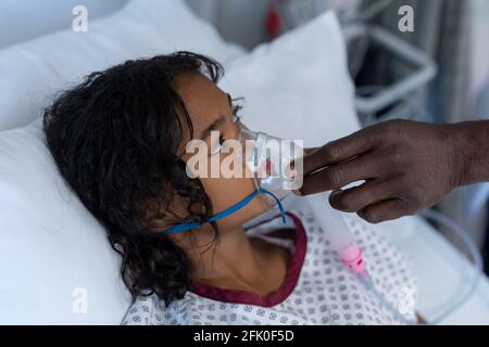 Main d'un médecin de sexe masculin mettant le ventilateur de masque d'oxygène sur malade fille de race mixte dans le lit d'hôpital Banque D'Images