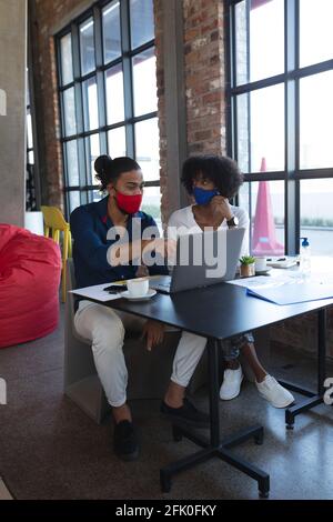 Femme afro-américaine et homme de race mixte portant des masques faciaux s'asseoir dans un café avec un ordinateur portable Banque D'Images