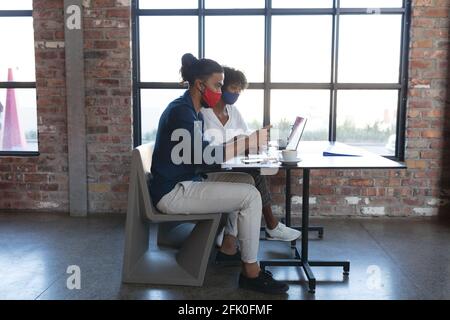 Femme afro-américaine et homme de race mixte portant des masques faciaux s'asseoir dans un café avec un ordinateur portable Banque D'Images