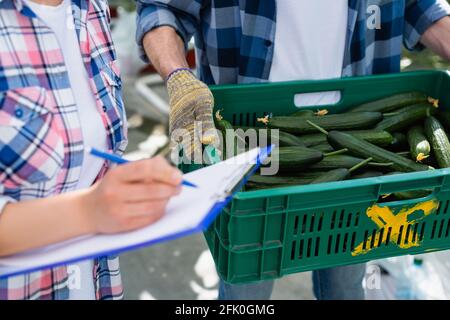 vue partielle sur le fermier avec boîte de concombres frais à proximité femme écrivant sur le presse-papiers sur un premier plan flou Banque D'Images