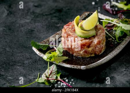 Tartare de thon avec salade verte, citron vert, avocat et sauce moutarde sur une plaque en céramique noire de style japonais sur fond de marbre noir. Restaurant Banque D'Images