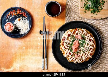 Crêpes au chou okonomiyaki maison au Japon, décorées d'oignons de printemps, de gingembre mariné, sauce mayonnaise sur plaque en céramique noire avec baguettes a Banque D'Images