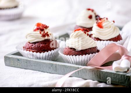 Petits gâteaux faits maison en velours rouge avec crème fouettée sur plateau en bois, serviette blanche avec ruban sur nappe blanche. Banque D'Images