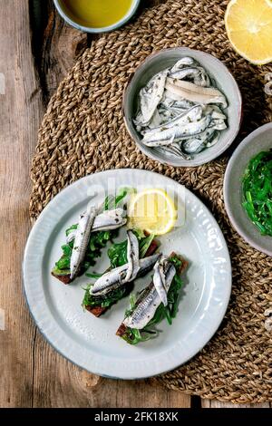 Hors-d'œuvre tapas Anchois marinés ou filet de sardines salade d'algues Wakame sur pain de seigle grillé, servi sur une assiette bleue avec citron sur un vieux backgro en bois Banque D'Images