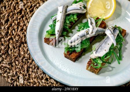 Hors-d'œuvre tapas Anchois marinés ou filet de sardines salade d'algues Wakame sur pain de seigle grillé, servi sur une assiette bleue avec citron sur la serviette de paille Banque D'Images