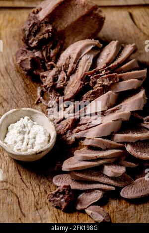 La langue de bœuf cuite au four est coupée en tranches et sert avec de la sauce au raifort sur une planche à découper en bois sur fond de bois. Gros plan. Banque D'Images