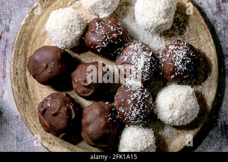 Boulettes de bonbons au chocolat à la noix de coco faites maison et légumes crus avec flocons de noix de coco dans une plaque en céramique sur fond de bois blanc. Pose à plat, gros plan Banque D'Images