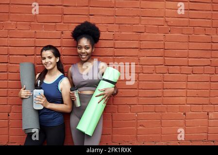 Deux jolies jeunes femmes en survêtements se tiennent contre un mur de briques dans la rue. Les amies sourient en tenant des tapis de yoga et des bouteilles d'eau. Banque D'Images