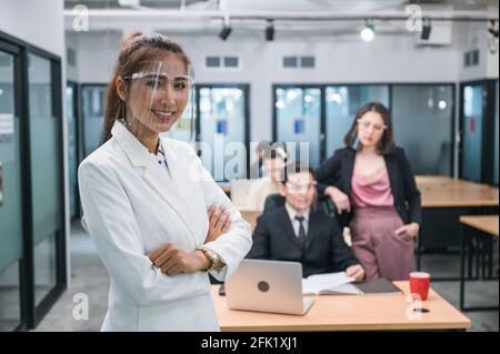 Femme asiatique confiante arborant un masque de protection souriant et croisée armes avec des collègues travaillant dans le bureau en arrière-plan Banque D'Images