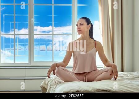 Grande fenêtre dans la chambre d'une jeune femme assise sur son lit dans des vêtements de maison roses. Banque D'Images