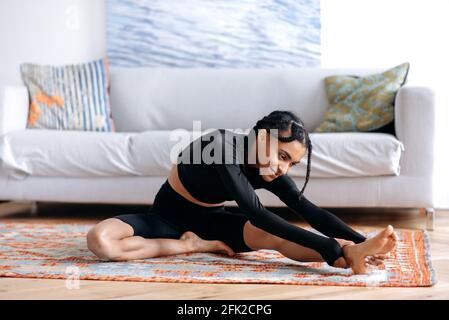 Afro-américaine fille de vêtements de sport, à la maison dans la salle de séjour sur la moquette, en étirant le dos, en étirant ses bras jusqu'au pied, est engagé dans la forme physique, mène un style de vie sain, sourit Banque D'Images