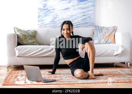 Jeune Afro-américaine à la figure sportive, dans des vêtements de sport, assis sur le sol de la salle de séjour, utilise un ordinateur portable, regarde la leçon vidéo de fitness, mène un mode de vie sain, regarde la caméra, sourit Banque D'Images