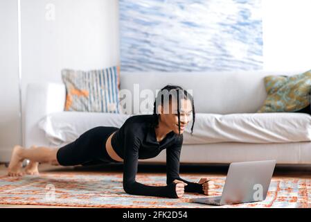 Belle jeune afro-américaine dans les vêtements de sport, faisant une planche sur le sol à la maison, à l'aide d'un ordinateur portable, regardant une vidéo de fitness ou des cours de yoga, menant un style de vie sain, souriant Banque D'Images