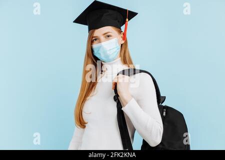 Bonne jeune femme diplômé en masque médical, étudiant en chapeau de remise de diplôme avec mallette sur fond bleu Banque D'Images