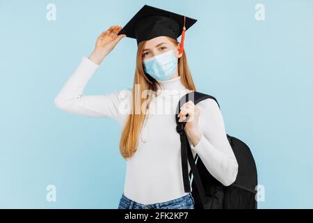 Bonne jeune femme diplômé en masque médical, étudiant en chapeau de remise de diplôme avec mallette sur fond bleu Banque D'Images