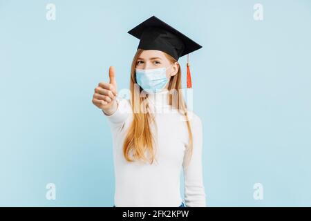 Bonne étudiante féminine diplômée en masque médical montrant le pouce vers le haut sur fond bleu Banque D'Images