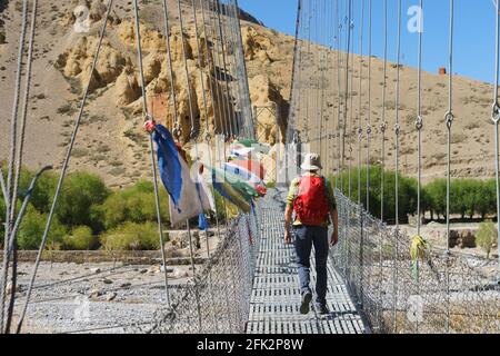 Trekker traversant un pont orné de drapeaux de prière tibétains à Chuksang, dans la région haute de Mustang, au Népal. Banque D'Images