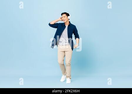 Portrait complet du corps d'un jeune homme asiatique souriant et charmant à l'écoute pour écouter de la musique avec un casque sans fil isolé dans un studio bleu clair arrière-plan Banque D'Images