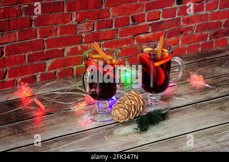 Deux verres de vin chaud avec des tranches de cannelle, d'anis, d'orange et de pomme se tiennent sur une table en bois contre un mur de brique avec une pile de l brûlant du nouvel an Banque D'Images