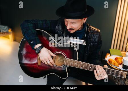 Image grand angle d'un homme d'âge moyen dans un chapeau assis sur un tabouret, portant une guitare à la maison. Il porte des vêtements décontractés et une veste en cuir sur le dessus. Banque D'Images