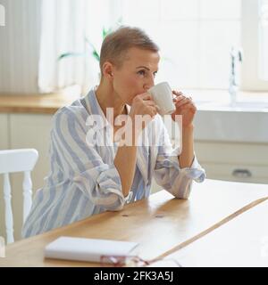 Boisson du matin. Vue latérale d'une belle femme de tête qui boit du café et pense à quelque chose pendant qu'elle est assise à une table en bois dans la cuisine, attention sélective sur la femme. Le mode de vie des personnes matures Banque D'Images