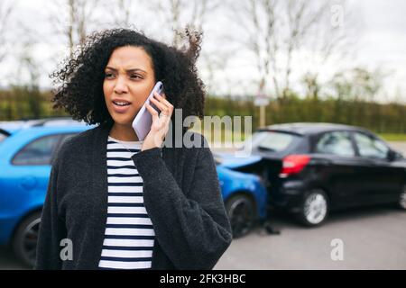 Jeune femme se tenant près d'une voiture endommagée après avoir signalé un accident de la route incident à une compagnie d'assurance utilisant un téléphone mobile Banque D'Images