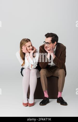 jeune couple en vêtements rétro souriant les uns aux autres assis dans une valise sur fond gris Banque D'Images