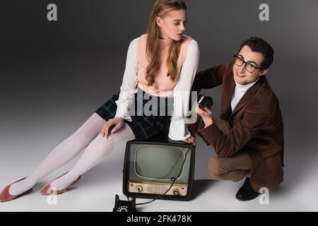 jolie femme assise sur une télé d'époque près d'un homme souriant tenant combiné sur fond gris Banque D'Images