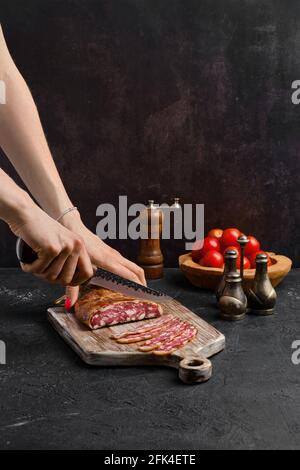 Main femelle avec couteau à trancher la saucisse de porc fumée dans bio boîtier sur planche à découper en bois Banque D'Images