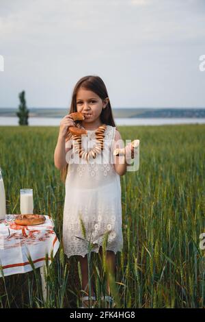 Portrait amusant de petite fille mignonne en robe blanche manger bagels avec lait dans le champ vert Banque D'Images