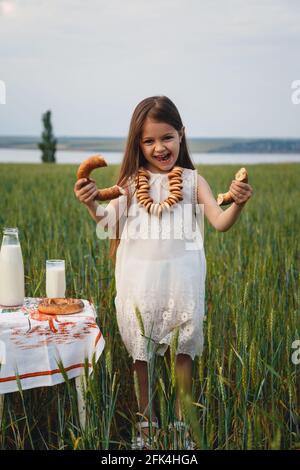 Portrait amusant de petite fille mignonne en robe blanche manger bagels avec lait dans le champ vert Banque D'Images