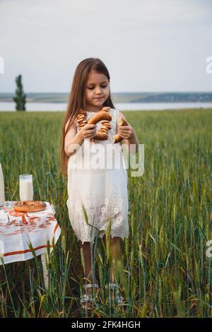 Jolie petite fille en robe blanche mangeant des bagels avec du lait dans le champ vert Banque D'Images