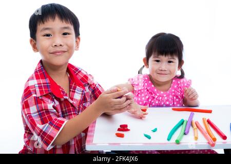 Petits enfants asiatiques jouant et créant des jouets à partir de la pâte de jeu sur la table. Garçon et fille souriant et regardant l'appareil photo, sur fond blanc. Renforcer Banque D'Images