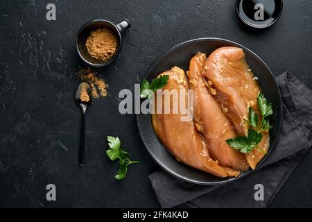 Filet de viande de poulet cru mariné dans de la sauce soya teriyaki, oignons, ail et poivre dans une assiette noire sur un fond d'ardoise foncé, de pierre ou de béton. Haut v Banque D'Images