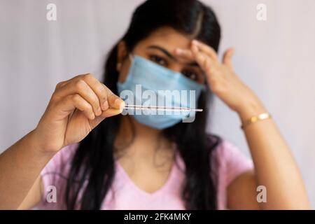 Accent sélectif sur un thermomètre analogique tenu à la main d'une femme indienne de taille moyenne, portant un masque nasal chirurgical et touchant le front, un confluent de soins de santé Banque D'Images