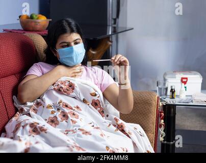 Une femme indienne de taille moyenne portant un masque nasal chirurgical vérifiant la température corporelle avec un thermomètre, allongé sur un canapé, concept de soins de santé Banque D'Images