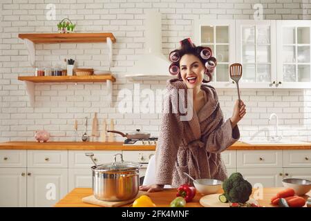 Femme en curlers et avec maquillage dans la cuisine dans un peignoir prépare un dîner romantique. Banque D'Images