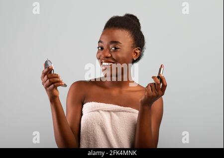 Femme afro-américaine faisant le maquillage quotidien, en utilisant le miroir et le rouge à lèvres sur fond gris studio. Banque D'Images